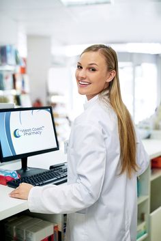 a woman in white lab coat standing next to a computer screen and smiling at the camera