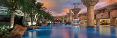 an indoor swimming pool surrounded by palm trees and water features large columns with lights on them