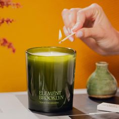 a person lighting a candle with a match in front of a green vase and orange wall