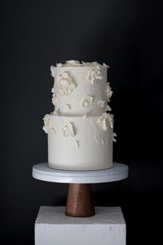 a three tiered white cake sitting on top of a wooden stand next to a black wall