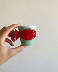 a hand holding a cup with a red fish on the inside and green bottom, in front of a white wall