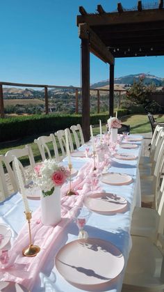 a long table set up with white and pink place settings for an outdoor wedding reception