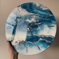 a hand holding a clock with blue and white marbled design on the face, in front of a beige wall