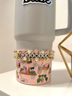 a pink coffee mug with the words babe on it next to a gold beaded bracelet