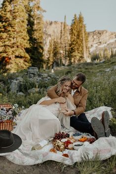 a man and woman are sitting on a blanket in the woods with food, drinks and snacks