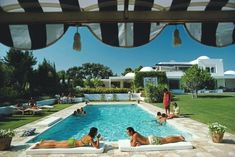 two women lounging on loungers in front of a swimming pool with black and white striped awnings