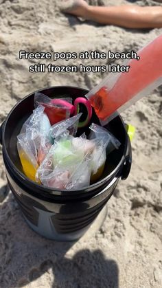 there is a bucket with plastic bags in it on the beach and someone's feet in the sand