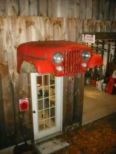 an old red jeep parked in front of a wooden wall with doors and window frames