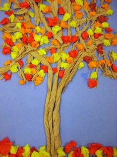 a tree made out of paper and colored tissue paper with leaves on the branches, sitting in front of a blue wall