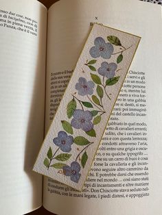 an open book with flowers and leaves on the page, next to it is a needle - embroidered bookmark