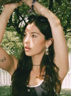 a beautiful young lady holding onto a tree branch with her hands on her head and looking off to the side