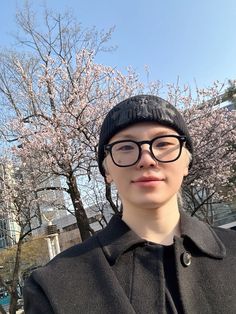 a woman wearing glasses and a black hat standing in front of trees with pink flowers