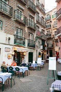 people are sitting at tables in an alleyway with balconies on the buildings