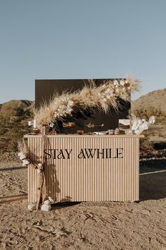 a sign that says stay awhile on it in the desert with dry grass growing out of it