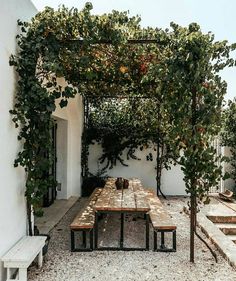 an outdoor table with benches and trees on the side of a white building in front of it