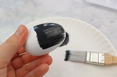 a hand holding a paintbrush next to an egg on a white plate with a black brush