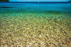 the water is crystal green and clear with rocks in it's foreground, as well as small sailboats on the horizon