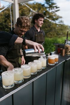 two men are making drinks at an outdoor bar, one is pouring something from a shaker