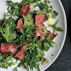 a white plate topped with meat and green leafy salad on top of a table