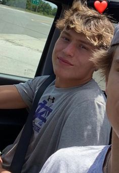 two boys sitting in the back seat of a car, one with a red heart on his head