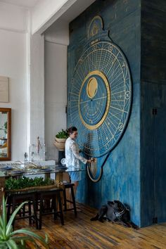 a woman standing in front of a blue wall with a large clock on it's side