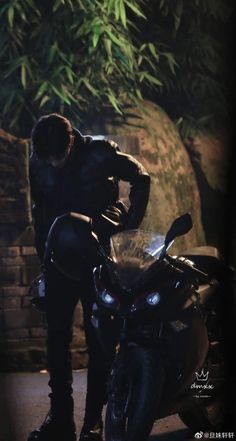 a man standing next to a motorcycle in front of a bushy area at night