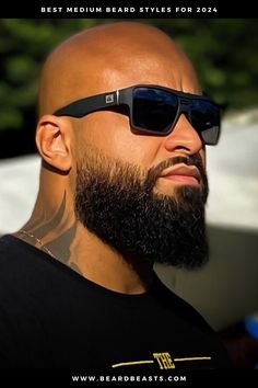 A man with a bold, medium-length beard is shown in profile, highlighting the fullness and well-groomed texture of his facial hair. His head is completely shaved, creating a striking contrast between the smooth scalp and the thick beard. He wears dark, stylish sunglasses and a black T-shirt, enhancing his confident and modern appearance. The beard is meticulously shaped, with clean lines that emphasize the strong jawline. Bold And Beard Style, Taper Beard For Men, Black Men Beard Style Ideas, Bald Men Beard Style, Bald Men With Beards Style, Barber Techniques, Mens Beard Styles