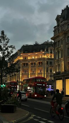 two red double decker buses driving down a street next to tall buildings at night time