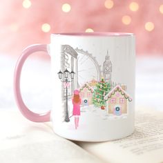 a pink and white coffee mug sitting on top of an open book next to a christmas tree