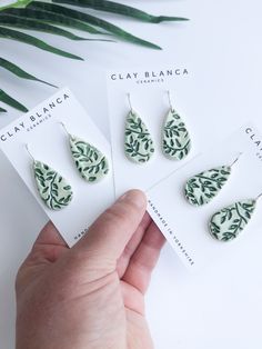 three pairs of green and white leaf print earrings on top of each other, one being held by a person's hand