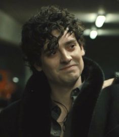 a man with curly hair wearing a black coat and looking at the camera while standing in a dark room