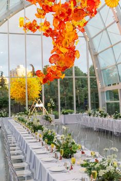 a long table is set up in front of a large glass window with flowers hanging from the ceiling