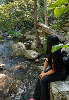 a woman sitting on top of a large rock next to a river in the forest