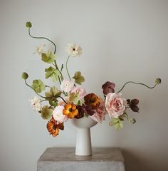 an arrangement of flowers in a white vase on a gray table against a white wall