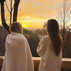 two women dressed in robes are standing on a balcony and looking at the sunset while holding coffee cups