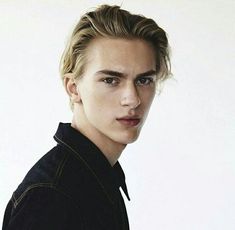 a man with blonde hair wearing a black shirt and looking at the camera while standing in front of a white wall