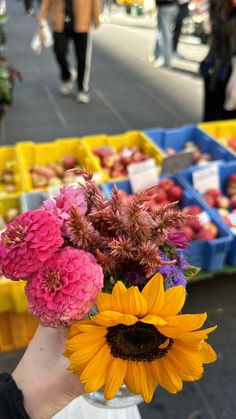 a person holding a vase filled with flowers on top of a street next to boxes of fruit