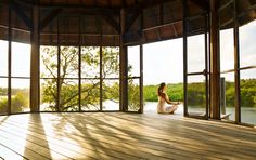 a woman sitting on a wooden floor looking out an open window at the water and trees