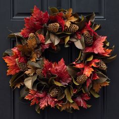 a wreath with leaves and pine cones hanging on a black front door, decorated with autumn foliage