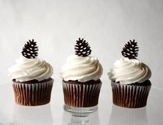 three chocolate cupcakes with white frosting and pine cones on top, sitting on a glass plate