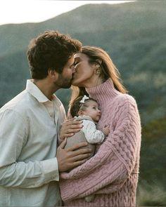 a man and woman kissing each other while holding a small child in their arms with mountains in the background