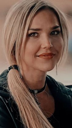 a woman with long blonde hair wearing a black leather jacket and silver necklace smiling at the camera