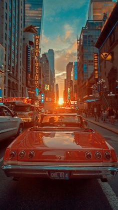 an old red car driving down a street next to tall buildings and traffic lights at sunset