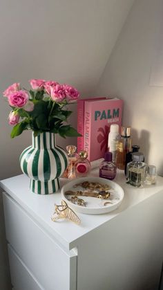 a vase with pink flowers on top of a white dresser next to perfume bottles and other items