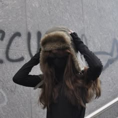 a woman with long hair wearing a fur hat and black gloves, standing in front of graffiti on a wall