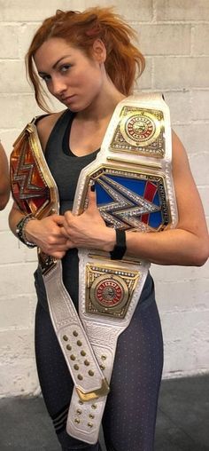 a woman in tight pants holding up two different colored wrestling belts on her chest and arm