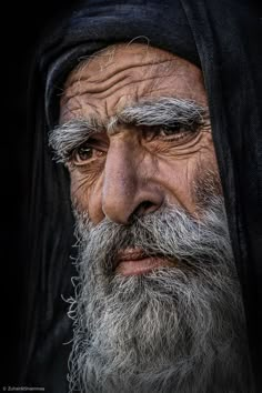 an old man with grey hair and beard wearing a black headdress is looking at the camera