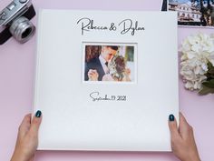 two hands holding a wedding album on a pink table with flowers and pictures in the background