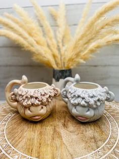 two ceramic sheep mugs sitting on top of a wooden table next to some dry grass