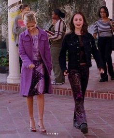 two women walking down the sidewalk in front of some people and one is wearing a purple coat